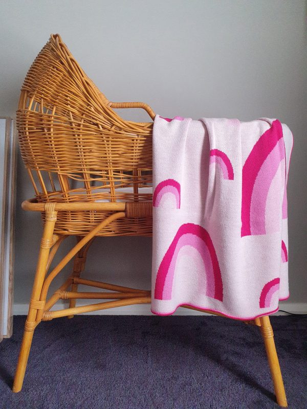 Merino wool Rainbow Baby Blanket in pink shades, draped over a rattan bassinet