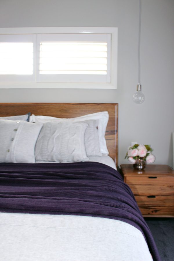 Bobbi in Currant Merino Wool Blanket draped over a wooden bed, paired with light grey knitted cushions, a matching side table, and peony flowers.