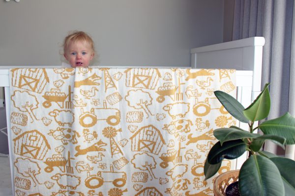 On The Farm Cot Blanket in Mustard and White, fully reversible. Close-up draped over the edge of a cot, showing half of the whole blanket with a toddler peeking over.