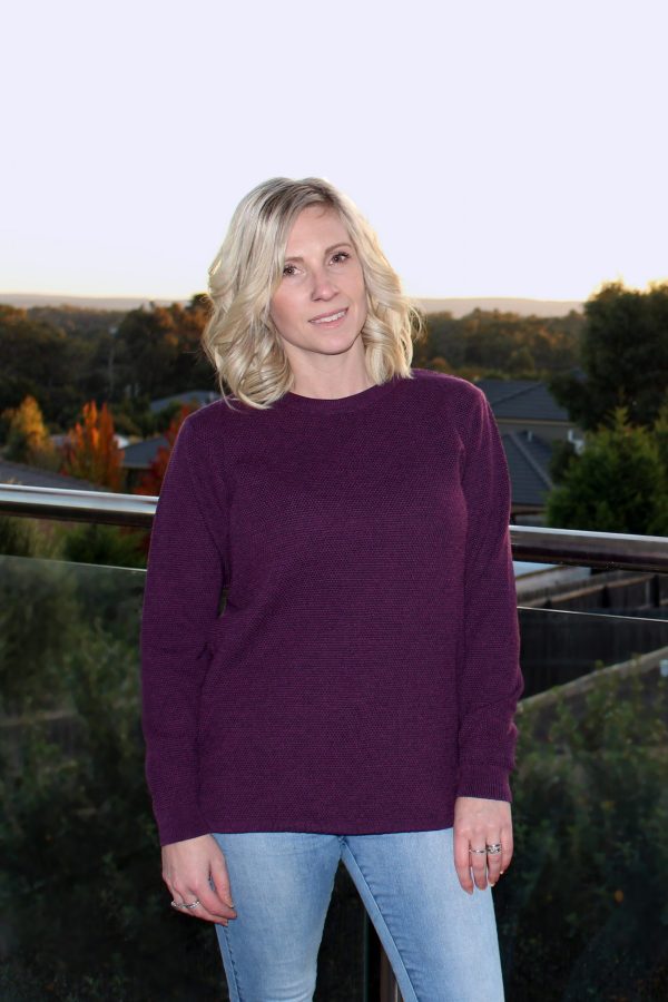 A lady standing on a balcony at sunset overlooking the trees, wearing an Australian Made, Ballarat Gold merino wool moss crew jumper in Currant (purple)