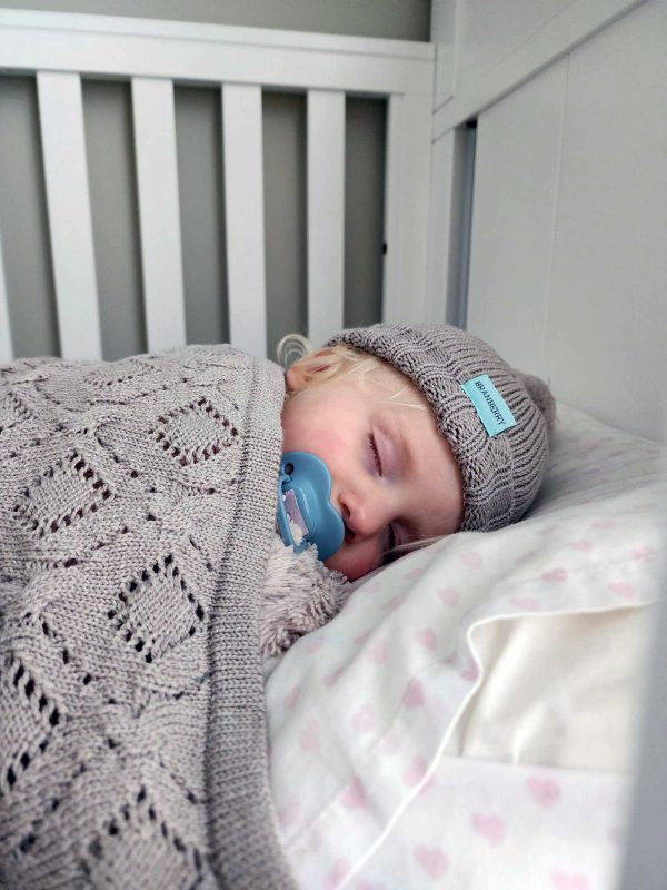 Branberry Heirloom Blanket in Cashew, a close-up view of a baby sleeping in a cot with the blanket and a matching Pure Wool Frankie Beanie.