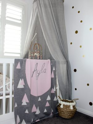 A Personalised Name Blanket with sporadic triangles with a big circle in the centre with a custom name, in a grey, pink and white colour combo draped over a cot. Made from Pure Australian Merino Wool and Made in Australia.