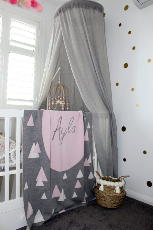 A Personalised Name Blanket with sporadic triangles with a big circle in the centre with a custom name, in a grey, pink and white colour combo draped over a cot. Made from Pure Australian Merino Wool and Made in Australia.