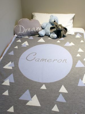A Personalised Name Blanket with sporadic triangles with a big circle in the centre with a custom name, in a grey, blue and white colour combo, placed on a bed. Made from Pure Australian Merino Wool and Made in Australia.