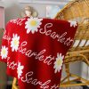 Australian Made, Merino Woollen Personalised Daisy Knitted Baby Blanket in Sangria Red, draped over the edge of a bassinet in a nursery. Features alternating an Daisy flower and a personalised name knitted in a script font.