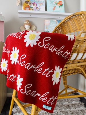 Australian Made, Merino Woollen Personalised Daisy Knitted Baby Blanket in Sangria Red, draped over the edge of a bassinet in a nursery. Features alternating an Daisy flower and a personalised name knitted in a script font.