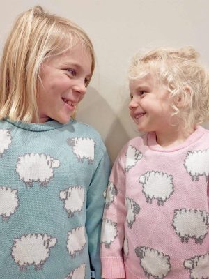 Two children standing indoors wearing a merino wool jumper with sheep patterns, smiling happily at each other.